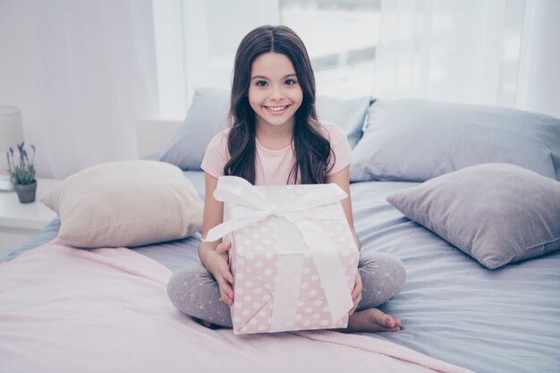 Linda garotinha sentada na cama com uma grande caixa de presente