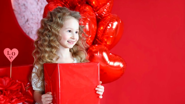 Linda garotinha sorri em um fundo vermelho com um balão em forma de coração e uma caixa de presente em suas mãos O conceito de presentes de amor Dia dos Namorados 14 de fevereiro 8 de março dia das mães