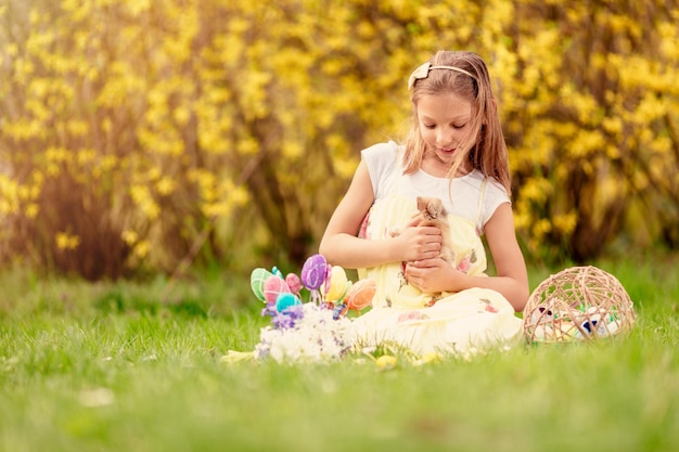 Linda garotinha sorridente segurando o coelhinho fofo e sentado na grama com ovos de Páscoa nas férias de primavera. Copie o espaço.