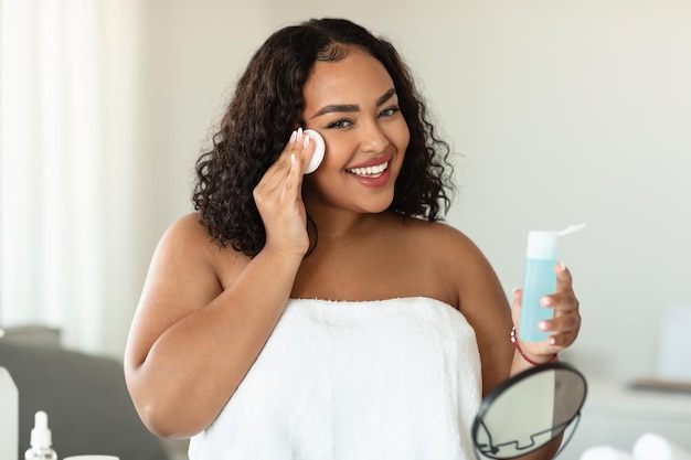 Foto linda gordinha preta limpando o rosto usando almofadas de algodão e produto de limpeza olhando para a câmera e sorrindo