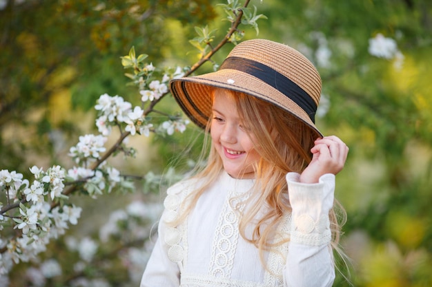 linda y graciosa niña rubia con un sombrero de paja cerca de un árbol floreciente en primavera riéndose