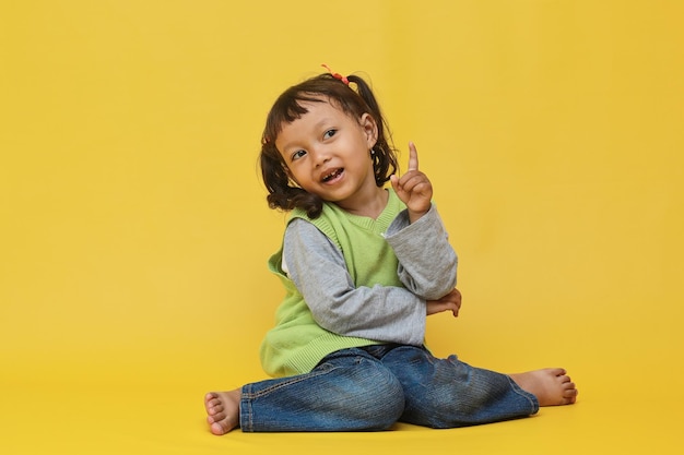 Linda y hermosa niña asiática sentada y apuntando hacia arriba aislada de fondo amarillo