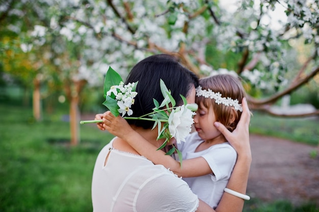 Linda hija y madre abrazándose en flor jardín de primavera Mujer feliz y niño, con vestido blanco al aire libre, se acerca la temporada de primavera. Concepto de vacaciones del día de las madres