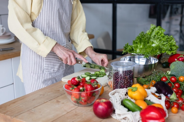 Linda idosa de cabelos grisalhos cozinha na cozinha aconchegante com legumes orgânicos frescos tomates repolho alface pepinos na mesa cozinhando salada de vegetais saudável comida saudável vida ativa