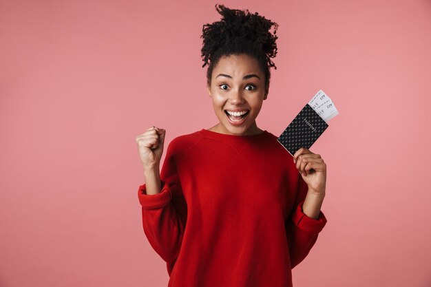 linda incrível feliz animado jovem africana posando isolado sobre a parede rosa, segurando o passaporte com ingressos.