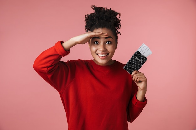 linda incrível feliz animado jovem africana posando isolado sobre a parede rosa, segurando o passaporte com ingressos.