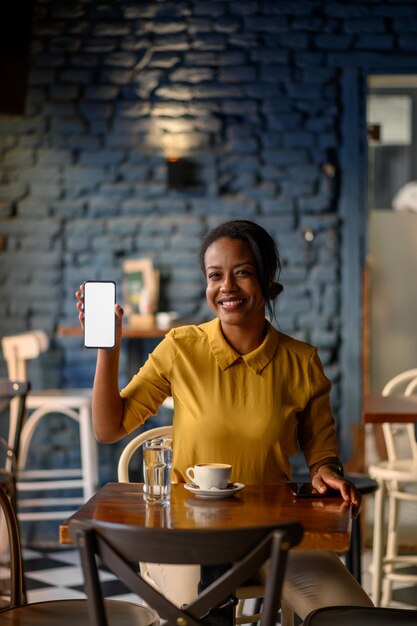 Foto linda jovem afro-americana mostrando um smartphone enquanto está sentado em um café