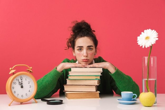 Linda jovem aluna entediada sentada à mesa isolada, estudando com os livros