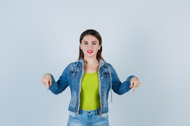 Foto linda jovem apontando para baixo em roupa jeans e olhando alegre, vista frontal.