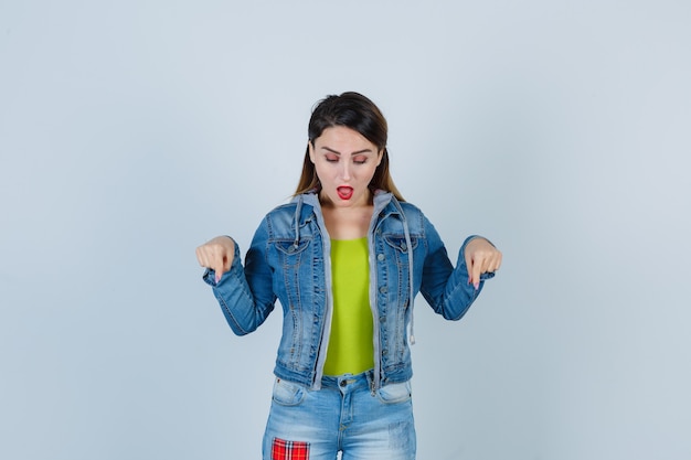 Linda jovem apontando para baixo em roupa jeans e olhando maravilhada, vista frontal.