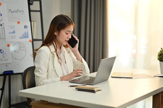 Linda jovem asiática CEO corporativa usando laptop e falando no celular em seu escritório