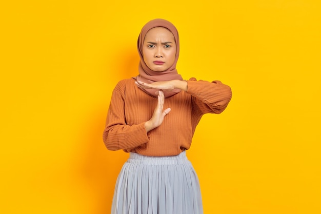 Linda jovem asiática de suéter marrom e hijab mostrando gesto de tempo com as mãos isoladas sobre fundo amarelo. Conceito religioso do islamismo de pessoas