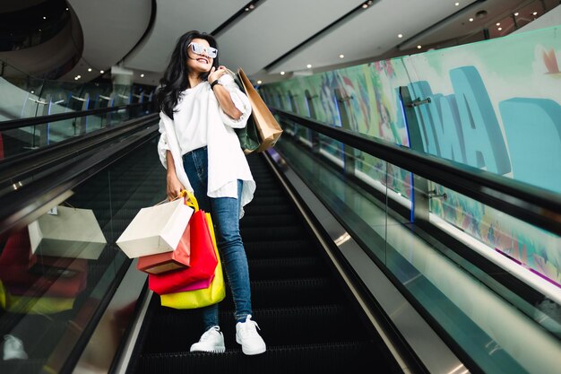 Linda jovem asiática segurando sacolas de compras e andando na escada rolante no shopping