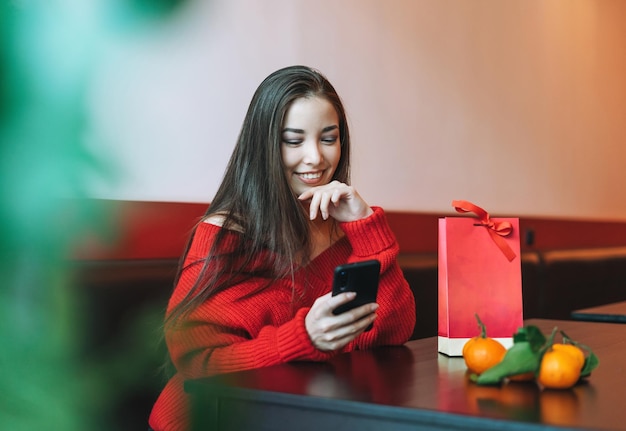 Linda jovem asiática sorridente em roupas vermelhas usando celular com saco de presente com mandarim no café