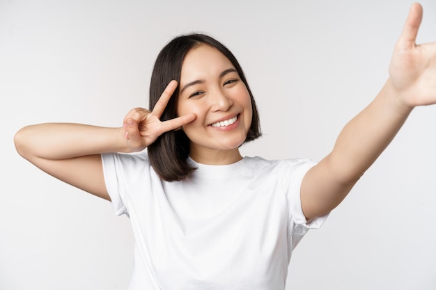 Linda jovem asiática tirando selfie posando com paz vsign sorrindo feliz tirando foto posando contra fundo branco