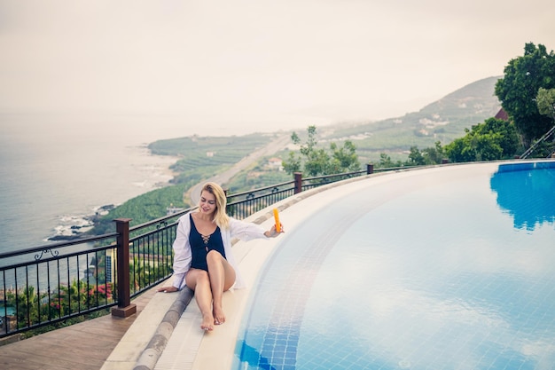 Linda jovem atraente senta-se perto de uma grande piscina com vista para o mar conceito de férias garota em um maiô preto mancha sua pele com protetor solar