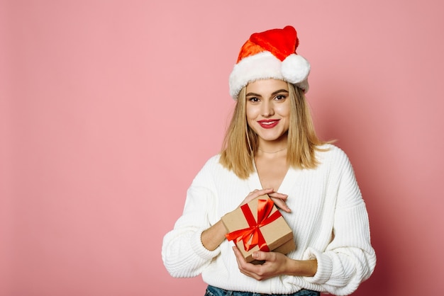 Linda jovem com chapéu de Papai Noel segurando um presente de Natal, sorrindo e olhando para a câmera