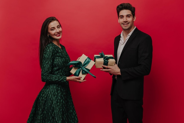 Linda jovem com penteado reto escuro e vestido verde e jovem bonito de terno preto de camisa branca segurando caixas de presente e olhando para a câmera contra fundo vermelho