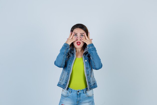 Foto linda jovem com roupa de jeans, olhando por entre os dedos e parecendo chocada, vista frontal.