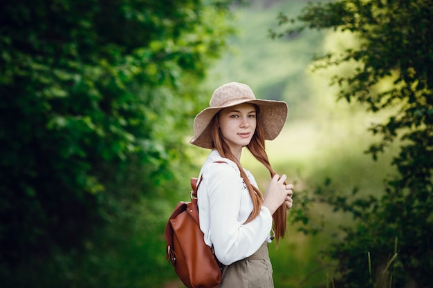 Linda jovem de cabelo ruivo, com um chapéu e uma mochila na orla da floresta. Linda garota ruiva passeando