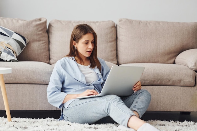 Linda jovem de jeans e camisa azul senta-se no chão com laptop dentro de casa em casa