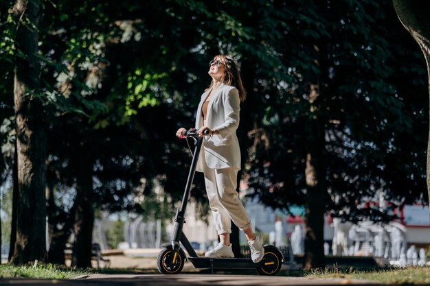 Linda jovem de óculos escuros e terno branco está de pé com sua scooter elétrica no parque da cidade