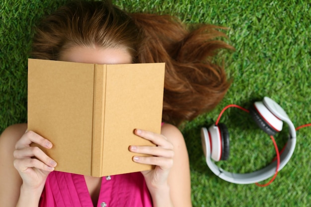 Linda jovem deitada na grama no parque segurando um livro. conceito de verão, lazer e educação. vista do topo