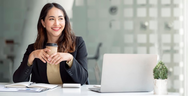 Linda jovem empresária asiática segurando uma xícara de café usando um tablet para trabalhar no laptop de calculadora de documentos contábeis colocado na mesa