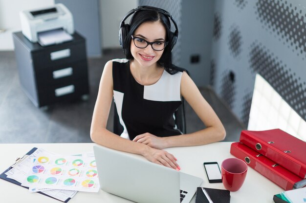 Linda jovem empresária de vestido preto, fones de ouvido e óculos, sentar à mesa e trabalhar no laptop