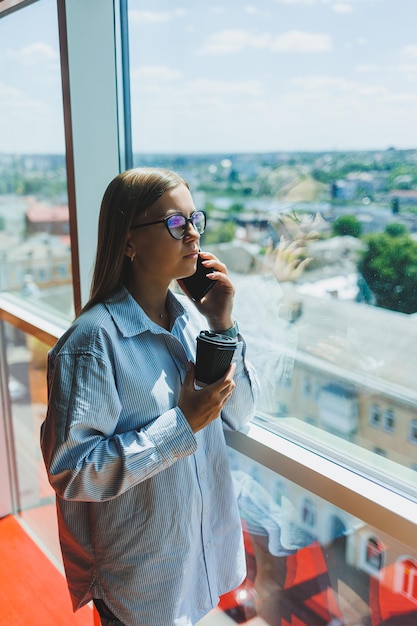 Linda jovem empresária falando no smartphone e trabalhando em um novo projeto de produtividade para a empresa, passando tempo livre no centro de coworking, apreciando a vista da janela