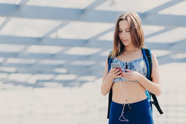 Linda jovem esportiva com aparência agradável tem cabelo escuro pele saudável vai para casa depois do treino lê mensagem no celular ouve música com fones de ouvido gosta de fim de semana ou dia de folga