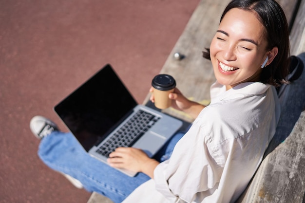 Linda jovem estudante asiática sentada com laptop no banco no parque bebendo café para viagem a