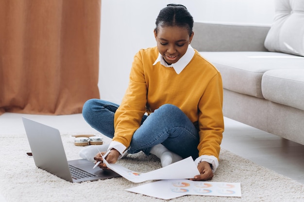 Linda jovem estudante universitária negra sentada no chão, bebendo café e fazendo projeto no laptop