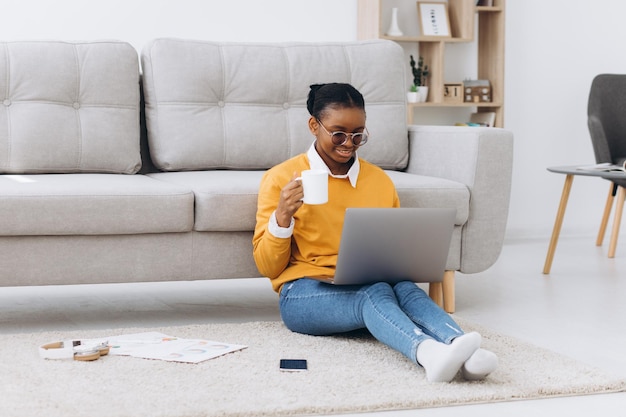 Linda jovem estudante universitária negra sentada no chão bebendo café e fazendo projeto no laptop