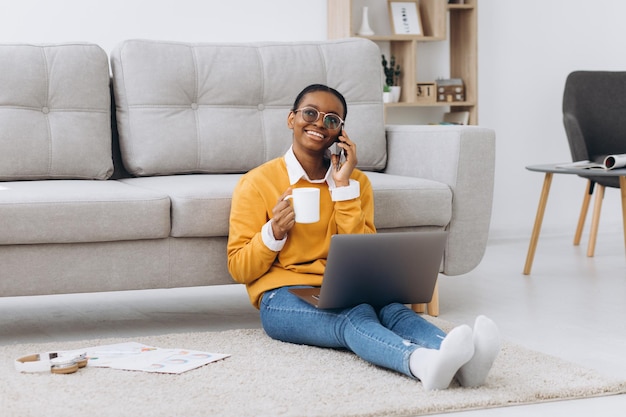 Linda jovem estudante universitária negra sentada no chão tomando café falando ao telefone e fazendo um projeto em um laptop