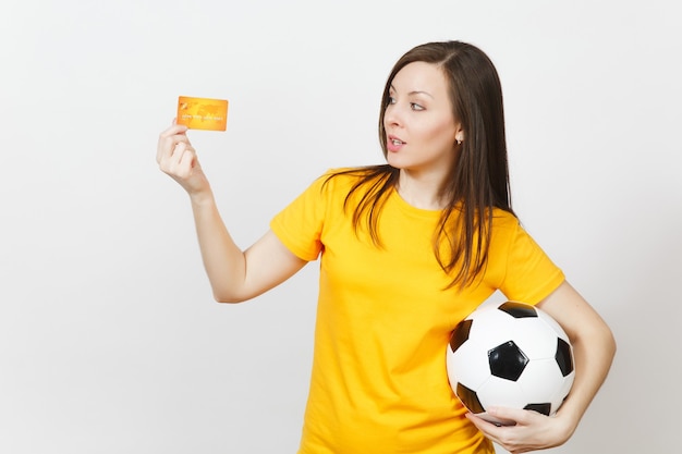 Linda jovem europeia alegre, fã de futebol ou jogador de uniforme amarelo, segurando uma bola de futebol de cartão de crédito isolada no fundo branco. Esporte, jogo de futebol, conceito de estilo de vida de excitação.