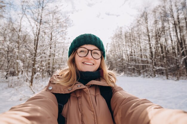 Linda jovem feliz com chapéu de malha de inverno verde leva selfie na floresta de inverno. Olhando para a câmera e sorrir. Viagem e conceito de vida ativa. Ao ar livre
