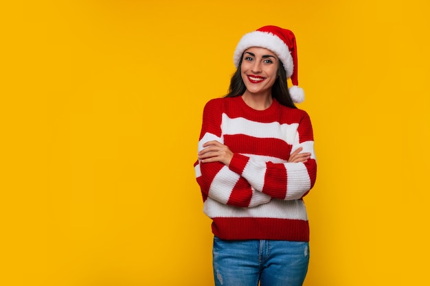 Linda jovem feliz com chapéu de Papai Noel e suéter de inverno posando isolada em um fundo amarelo na época do Natal