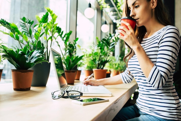 Linda jovem feliz usando laptop e bebendo café no café durante a hora do almoço