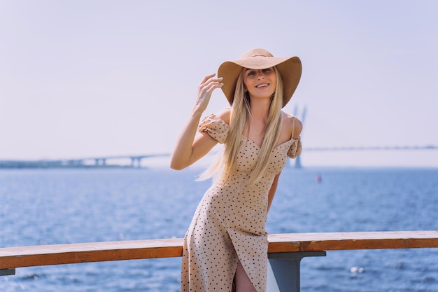 Linda jovem italiana linda em vestido bege fica no aterro da baía com chapéu de palha contra o mar e uma ponte enorme Sorrindo muito feminina andando no verão Férias de viagem de lazer