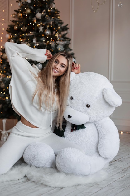 Linda jovem loira com um lindo sorriso branco como a neve vestindo uma roupa de moda branca com capuz senta-se ao lado de um ursinho de pelúcia perto da árvore de Natal em casa