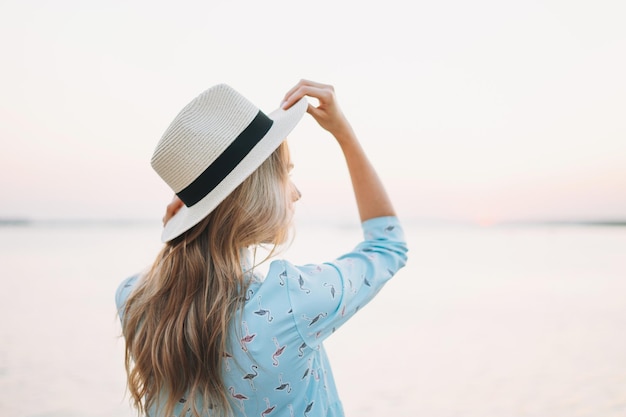 Linda jovem loira de vestido azul e chapéu de palha no cais no pôr do sol