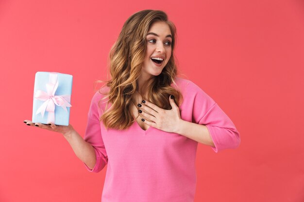 Foto linda jovem loira em pé isolada sobre uma parede rosa, mostrando uma caixa de presente