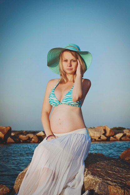 Linda jovem loira grávida elegante com um chapéu azul caminha na praia, beira-mar ao pôr do sol.