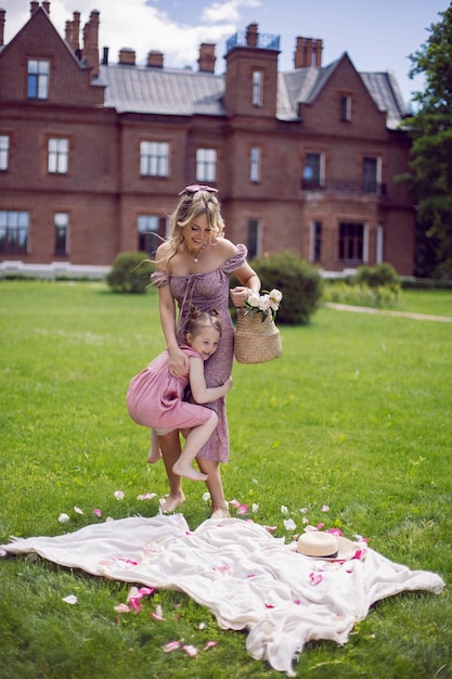 Linda jovem mãe com filha engraçada em vestidos rosa fica em uma manta em um prado verde na natureza com um piquenique no verão