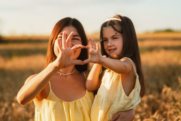 Linda jovem mãe e sua filha abraçando e fazendo coração com os dedos em um campo de trigo ao pôr do sol