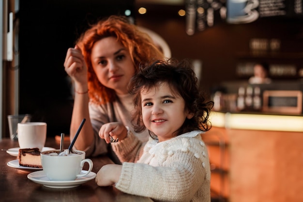 Linda jovem mãe ruiva com uma linda filha de cabelos encaracolados está sentada em um café aconchegante e bebendo um dia quente da escola das mães