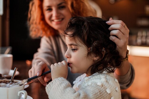 Linda jovem mãe ruiva com uma linda filha de cabelos encaracolados está sentada em um café aconchegante e bebendo um dia quente da escola das mães