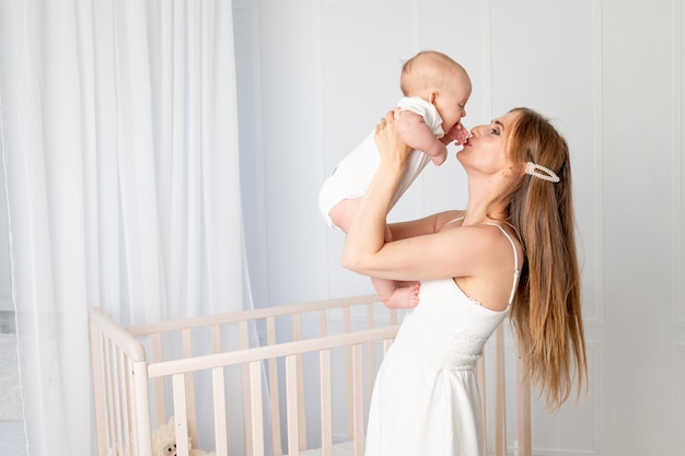 Linda jovem mãe segurando sua filha, abraçando e beijando-a no berçário em pé ao lado do berço, dia das mães, lugar para texto