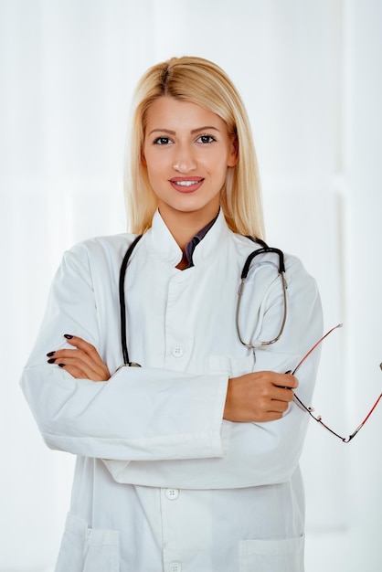 Linda jovem médica bonita em um jaleco branco em pé no escritório com um estetoscópio no pescoço. Olhando para a câmera.